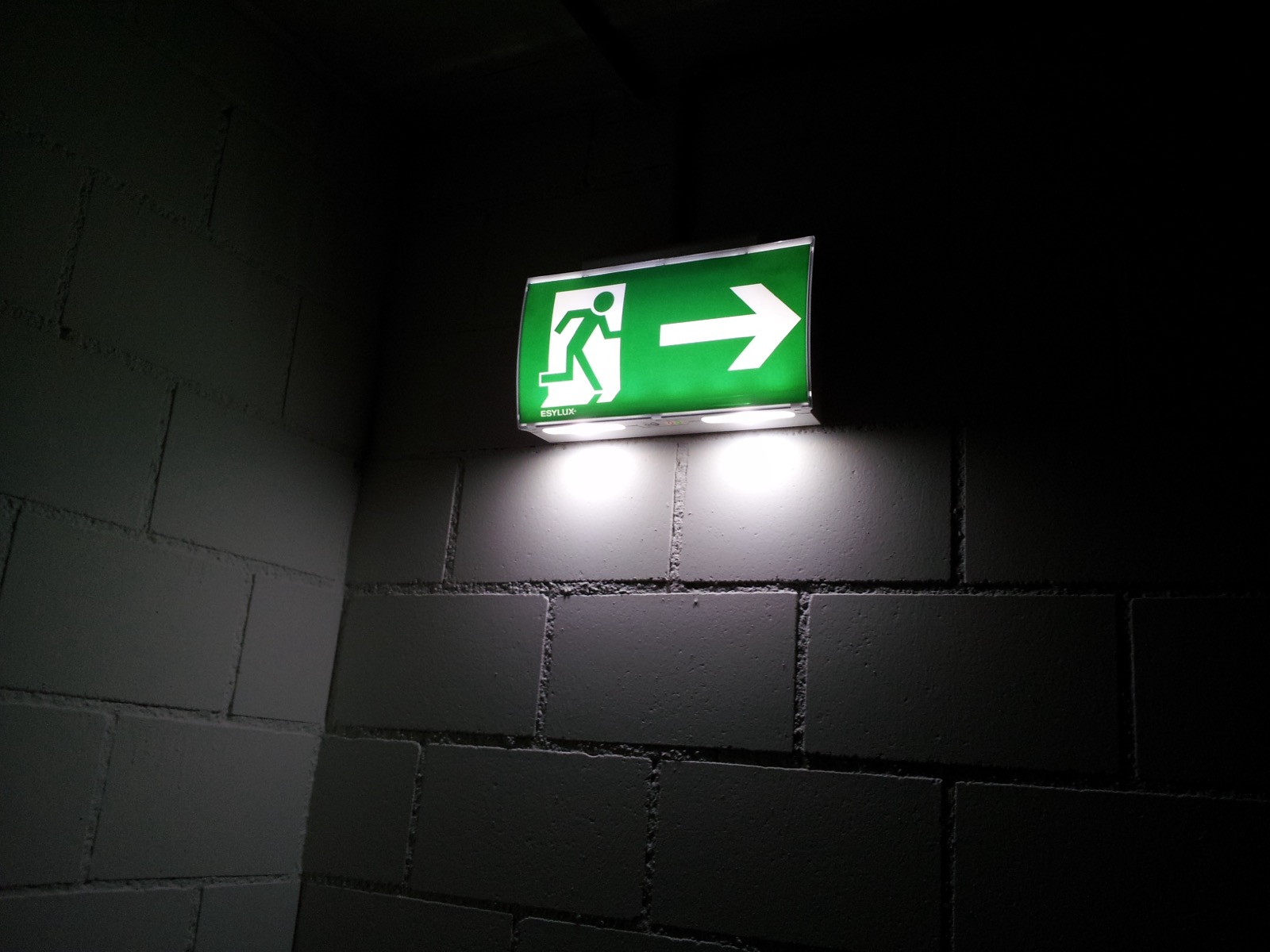 Illuminated green emergency exit sign with a running figure and arrow on a block wall in a dark corridor
