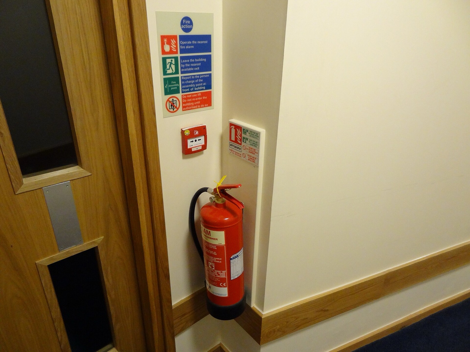 Fire extinguisher, manual call point and fire action notice on a corridor wall beside a timber door