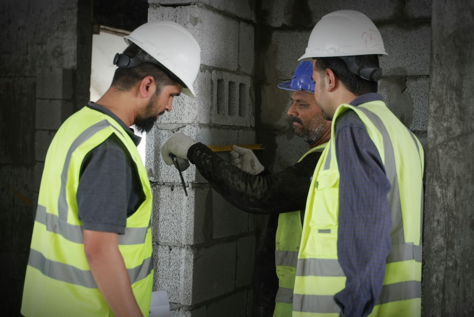 Three construction workers in hard hats and high-visibility vests measuring a block wall together